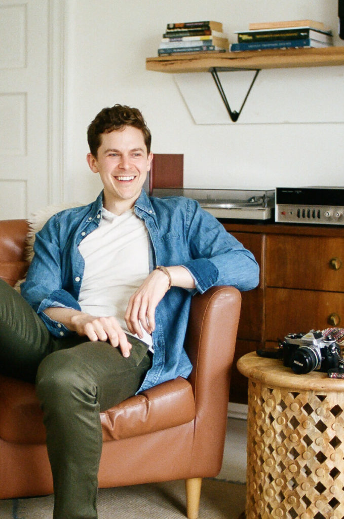 A Philly wedding photographer sits in an armchair smiling with a camera on a table beside him.