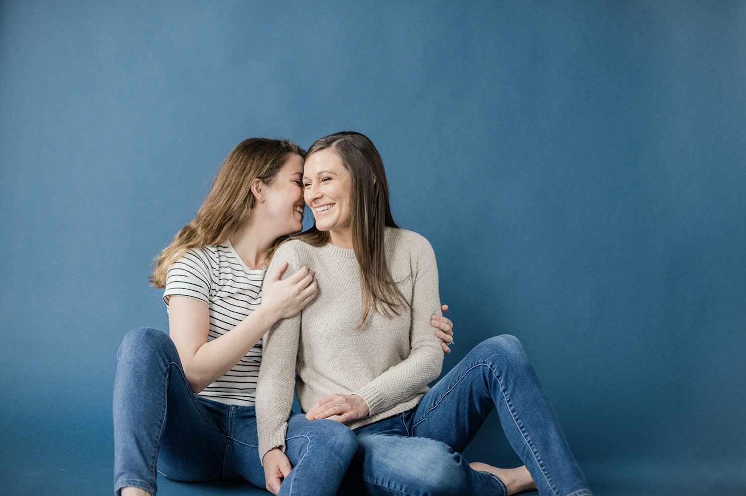 Woman whispering in partner's ear as she laughs during their engagement shoot where they both wear matching denim jeans and sit with a blue backdrop behind them, taken by Jason Moody Photography
