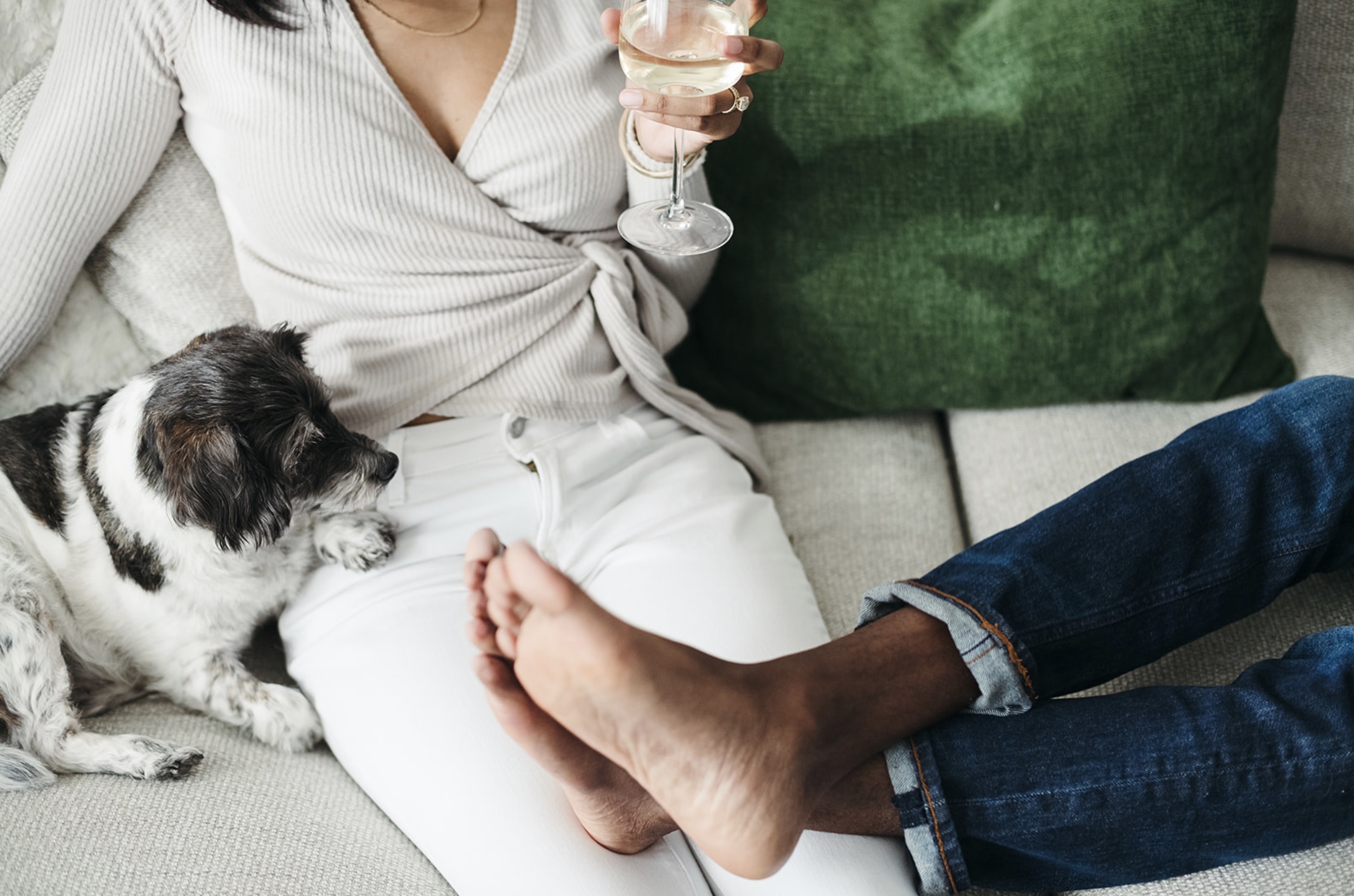 Close-up shot of girl sitting on the couch and holding a glass of champagne with the guy's feet on her lap and their dog beside her
