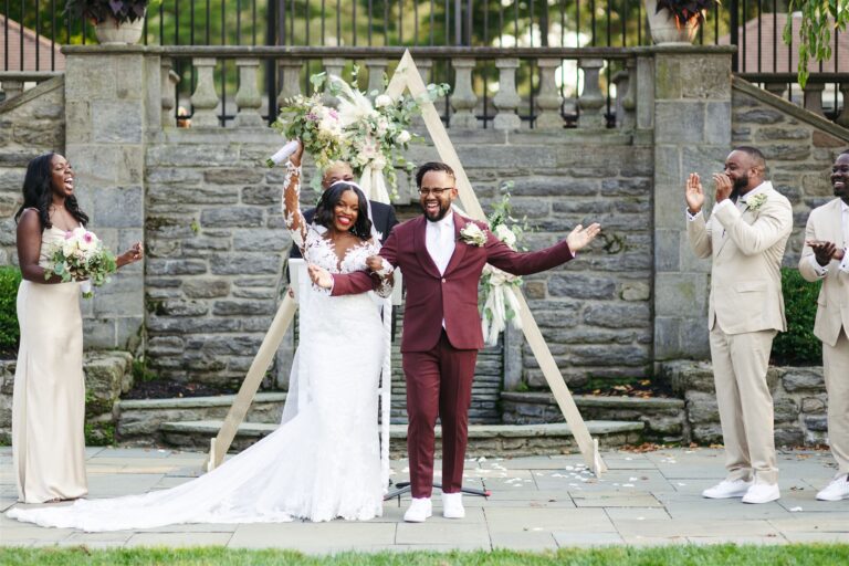 Why Is Wedding Photography so Expensive: Bride and groom raising their hands after they are pronounced husband and wife with their entourage applauding them, captured by Jason Moody Photography