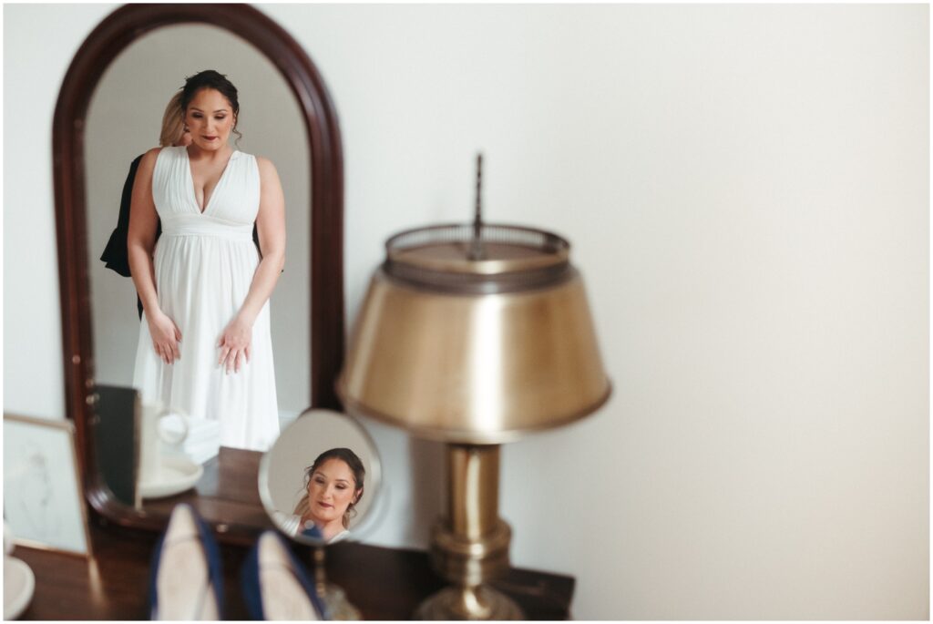 A woman puts on a white wedding dress while looking in a floor length mirror in her getting ready suite.
