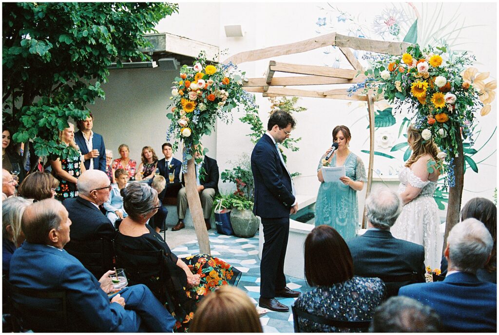 A couple stands under a floral arch in a courtyard wedding venue.