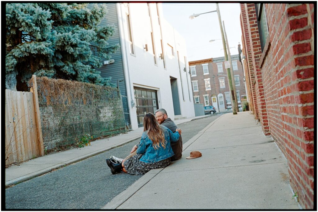 A couple sits on a city sidewalk in a Philadelphia engagement photo session.