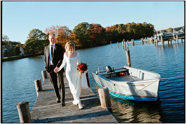 A couple holds hands and walks along a dock at their backyard wedding beside a lake.