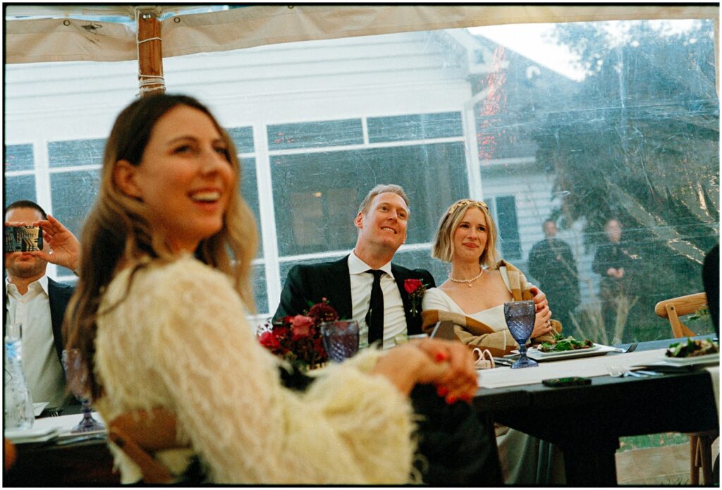 A bride and groom listen to speeches at a backyard wedding reception.