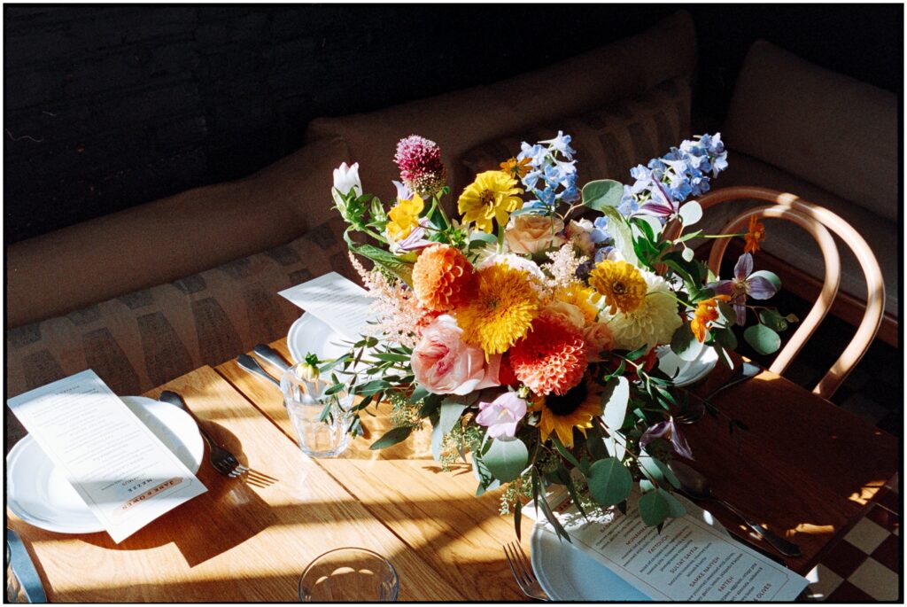 Bright flowers sit at the center of a wedding reception table in a patch of sun.