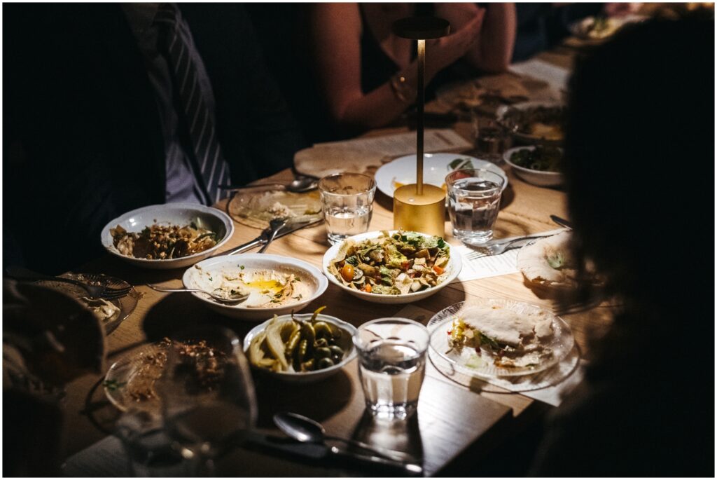 Plates of Mediterranean food sit clustered on a dimly lit table.