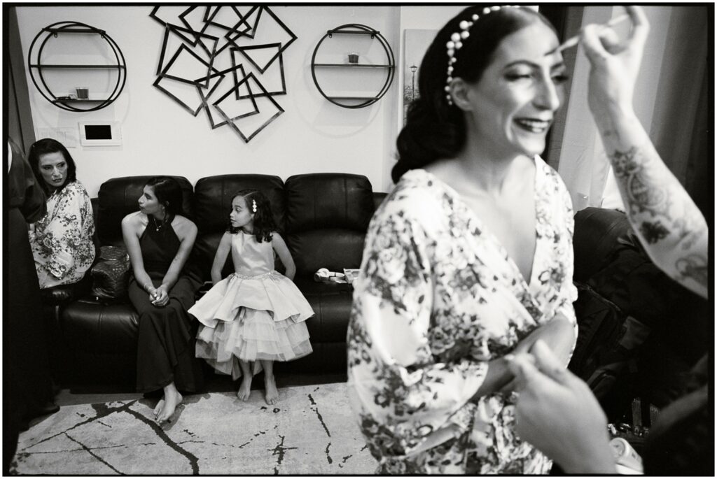 A makeup artist applies makeup to a bride while two children sit on a couch.