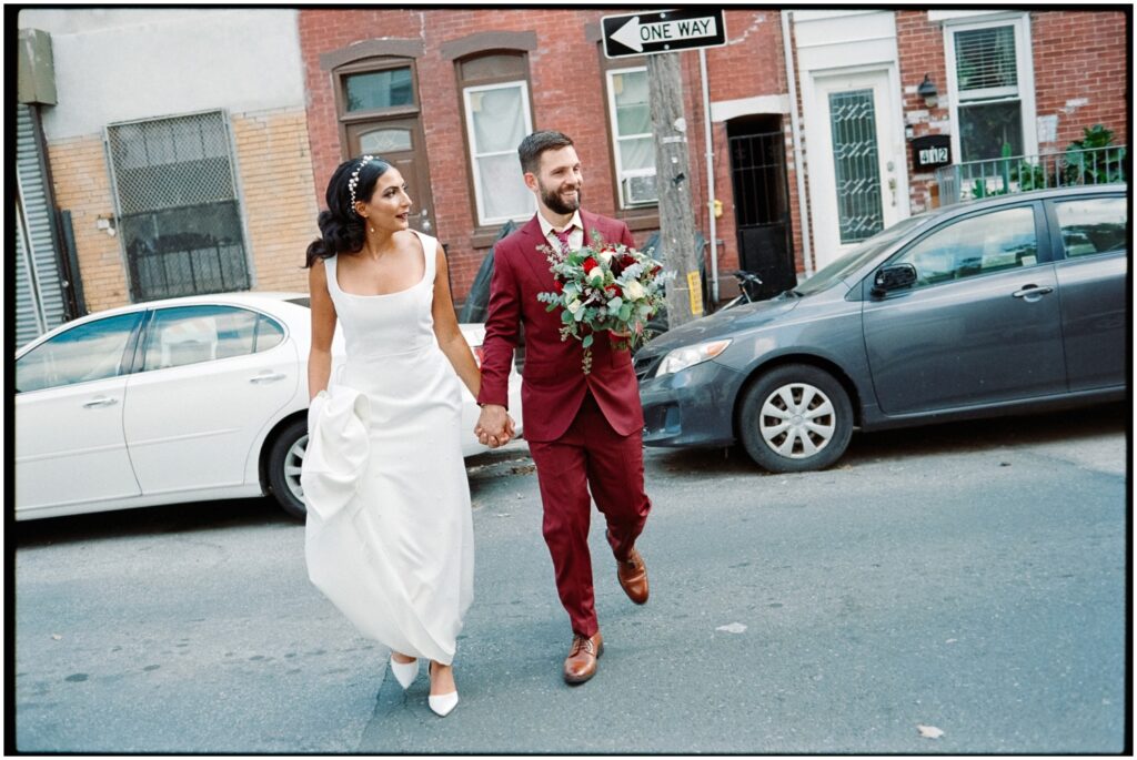 A groom carries a bridal bouquet as he crosses a street with a bride in film wedding photography.