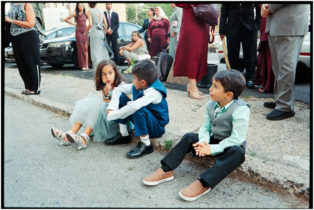 Children sit on a sidewalk as adults walk past.