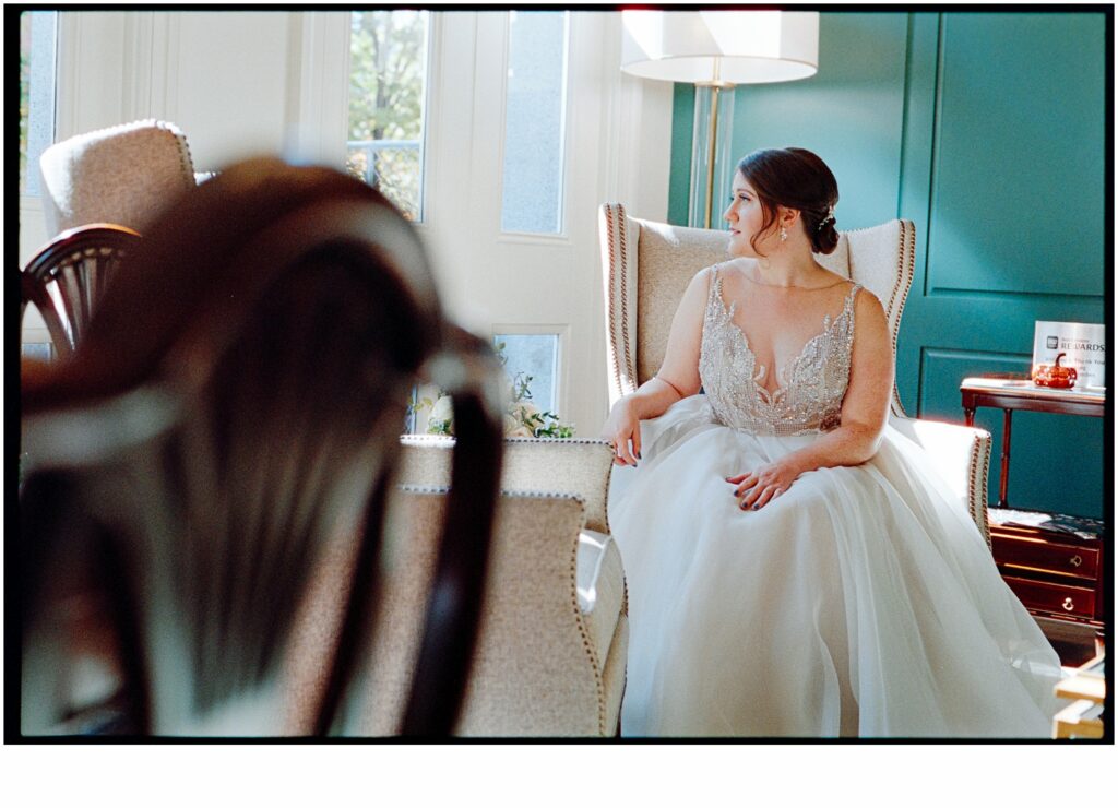 A bride looks over her shoulder seated in a white chair beside a tall window.