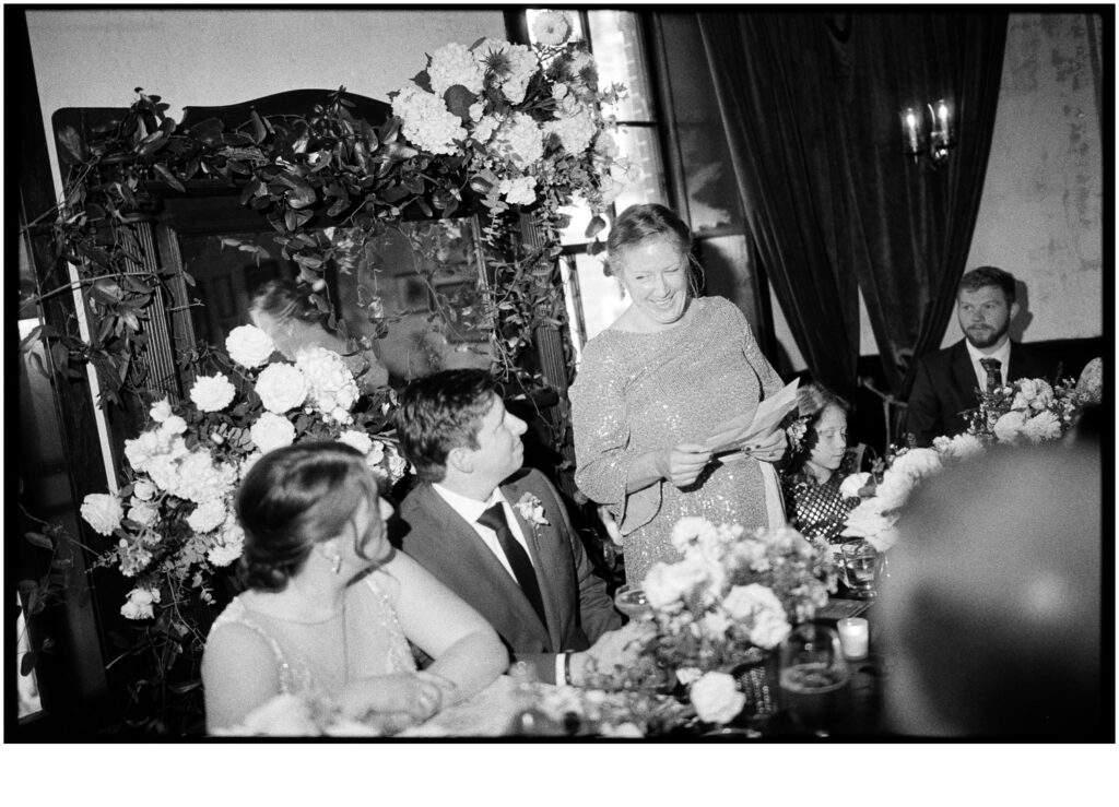 A wedding guest stands next to a bride and groom reading a toast from her phone.