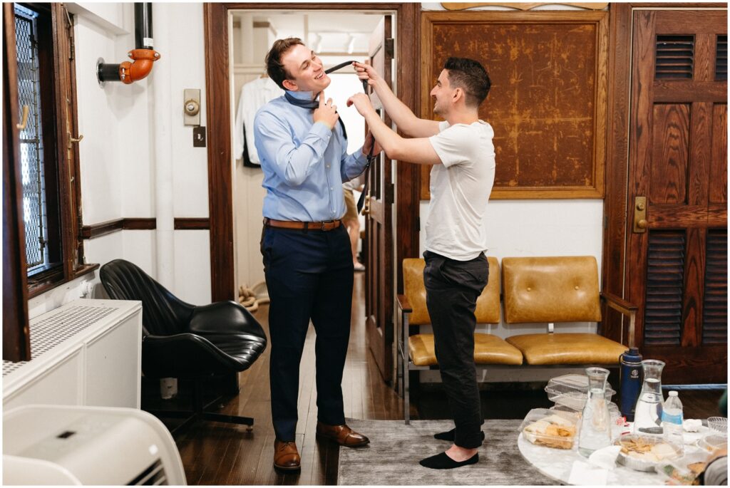 Two groomsmen tie a necktie in a getting ready suite and talk about how to have a small wedding