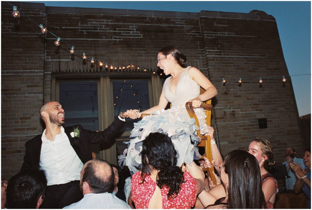 Wedding guests raise a bride and groom in chairs to dance the horah.