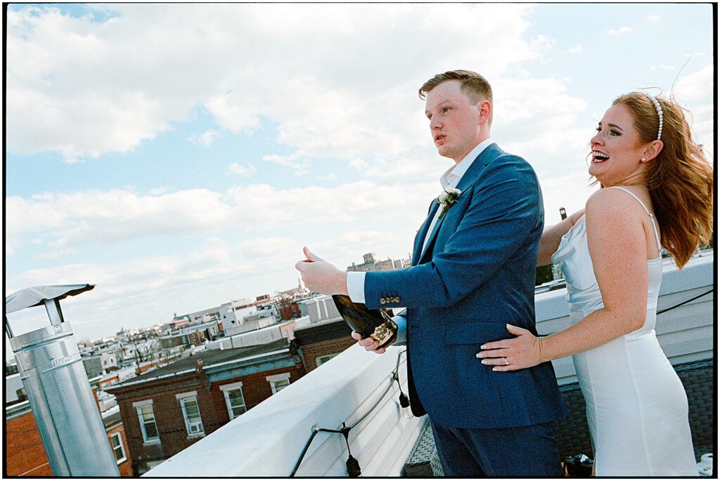A bride stands behind a groom popping champagne on a rooftop during their Philadelphia City hall wedding.