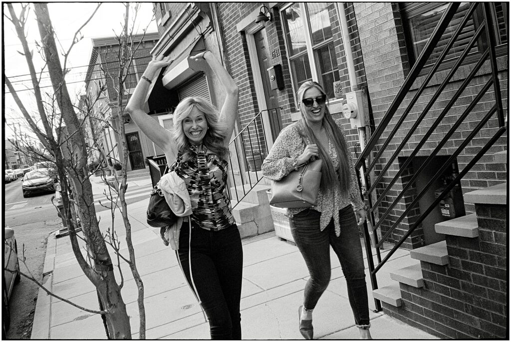 Two women walk down a Philadelphia street smiling at a wedding photographer.