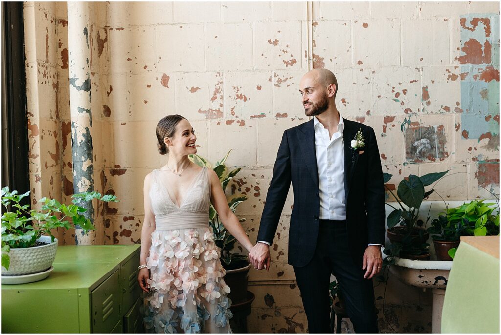 A bride and groom hold hands and smile at each other in a sunny room in the BOK Building.