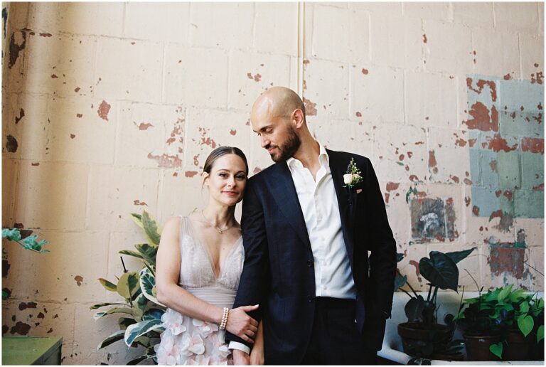 A bride in a blue and pink dress holds a groom's hands and leans against him.