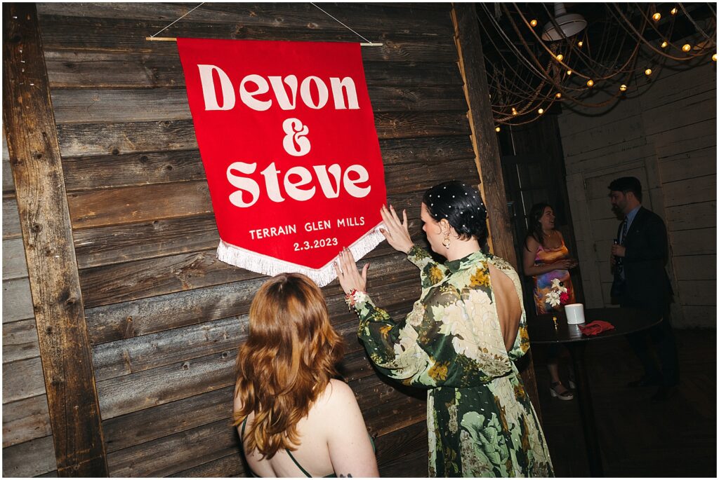 Two guests admire Devon and Steve's custom wedding sign hanging on the wall at Terrain.
