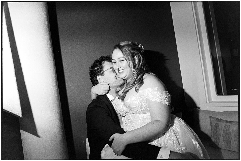 A bride sits on a groom's lap, and he kisses her cheek.