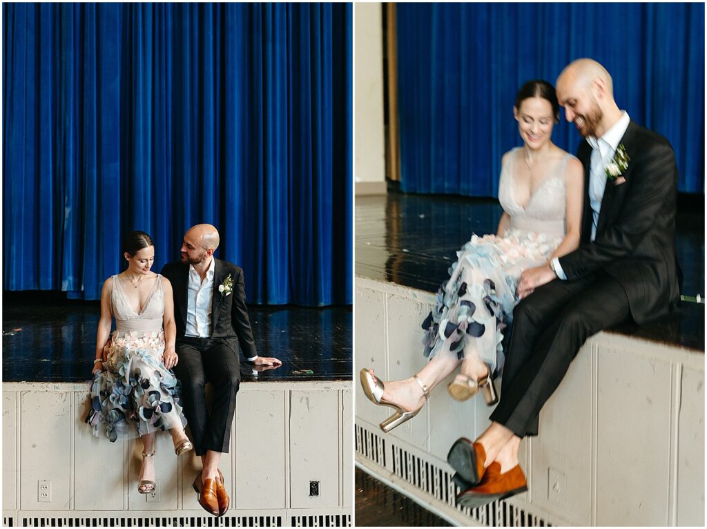 A bride and groom sit on the stage of an old auditorium with a blue curtain behind them.