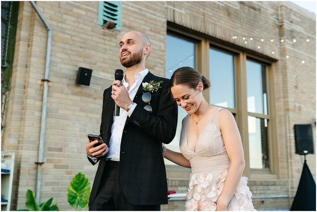 A bride laughs while a groom gives a wedding speech.