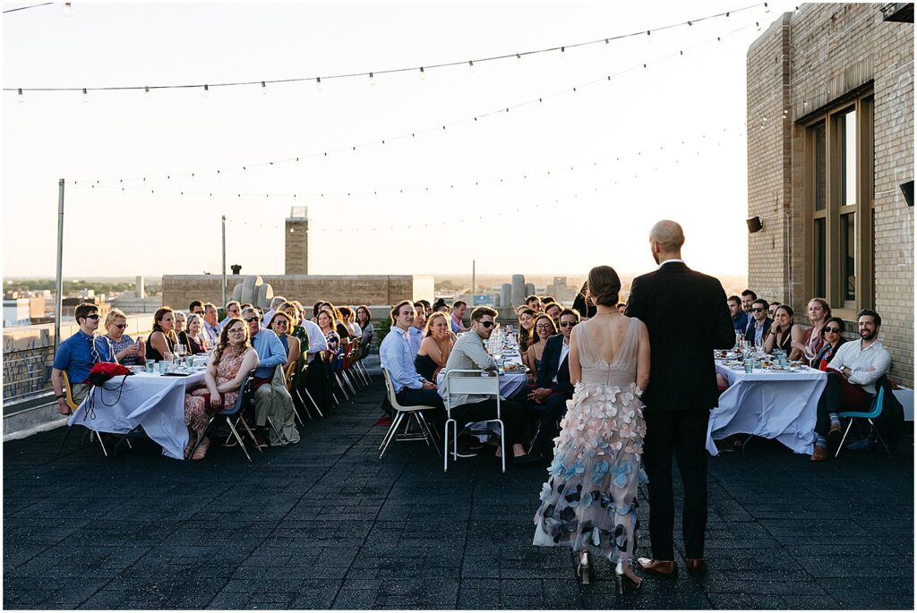 Wedding guests sit at long reception tables and listen to a groom's speech at a rooftop wedding at the BOK Building.