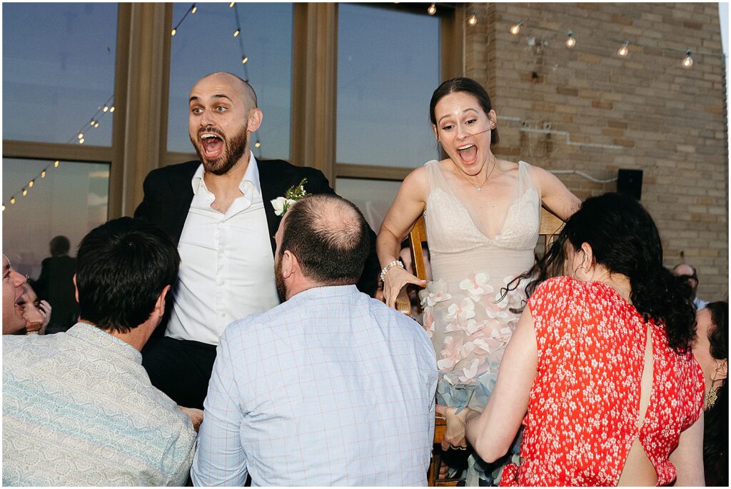 A bride and groom laugh as guests raise them for the horah.