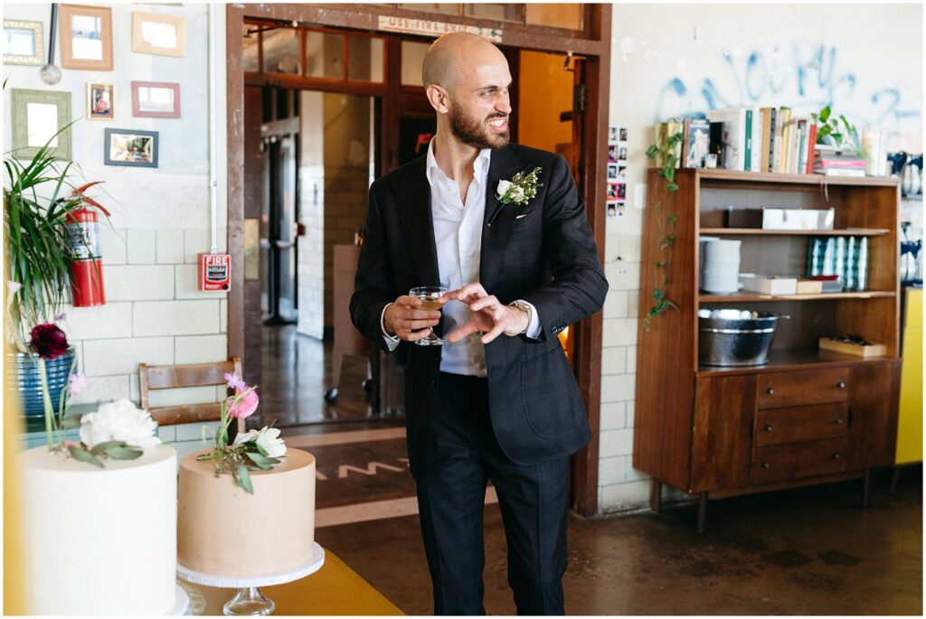 A groom holds a cocktail beside a table with two minimalist wedding cakes decorated with flowers.