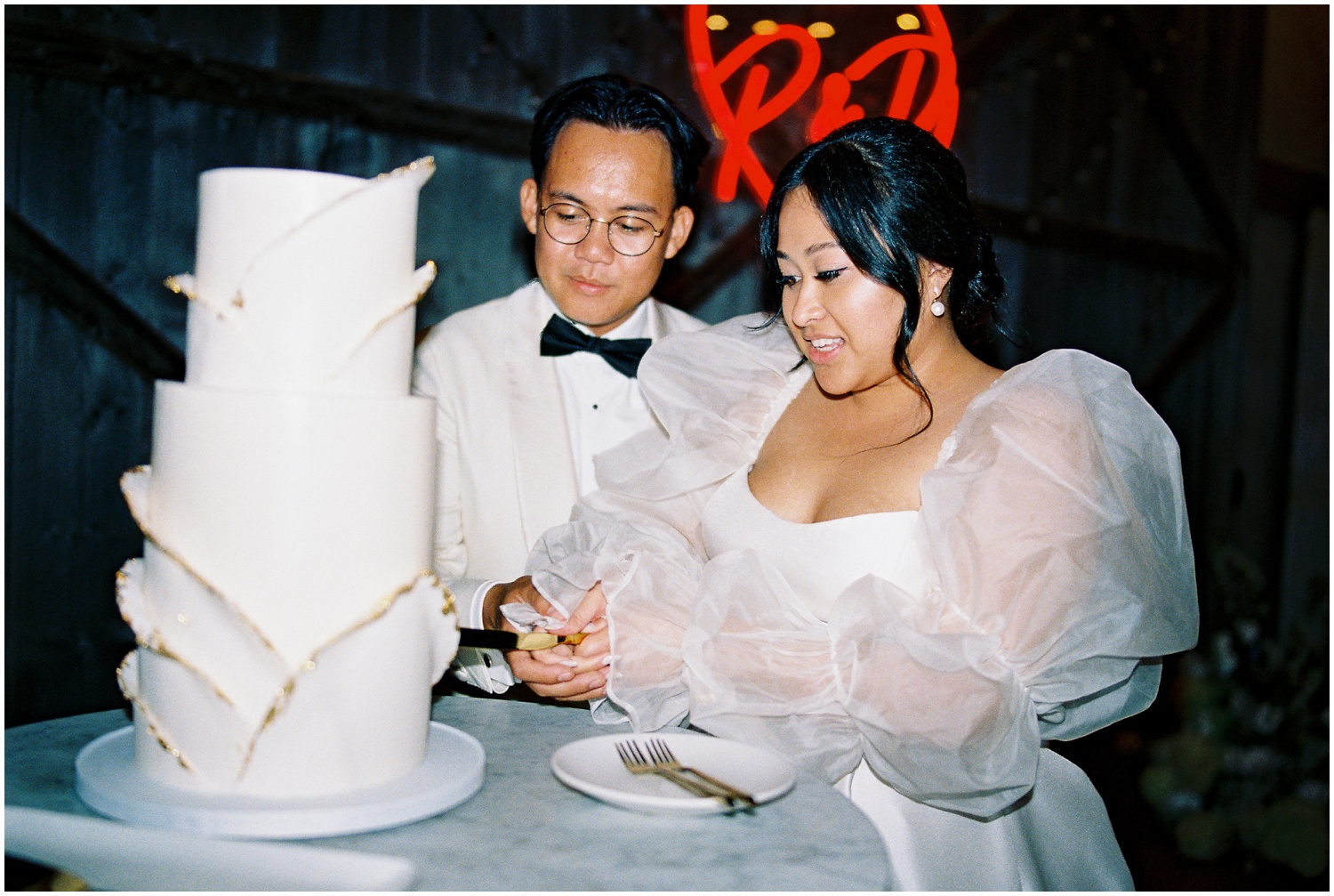 A bride and groom cut a wedding cake beneath a custom neon wedding sign.