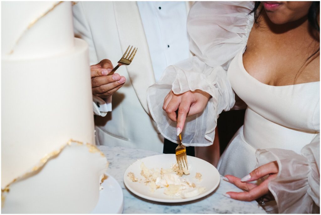 A couple lifts bites of wedding cake off a plate with gold forks.