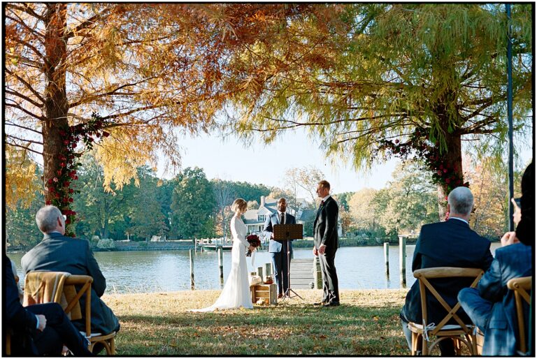 A bride and groom exchange vows at a lake house wedding with fall leaves in the background.