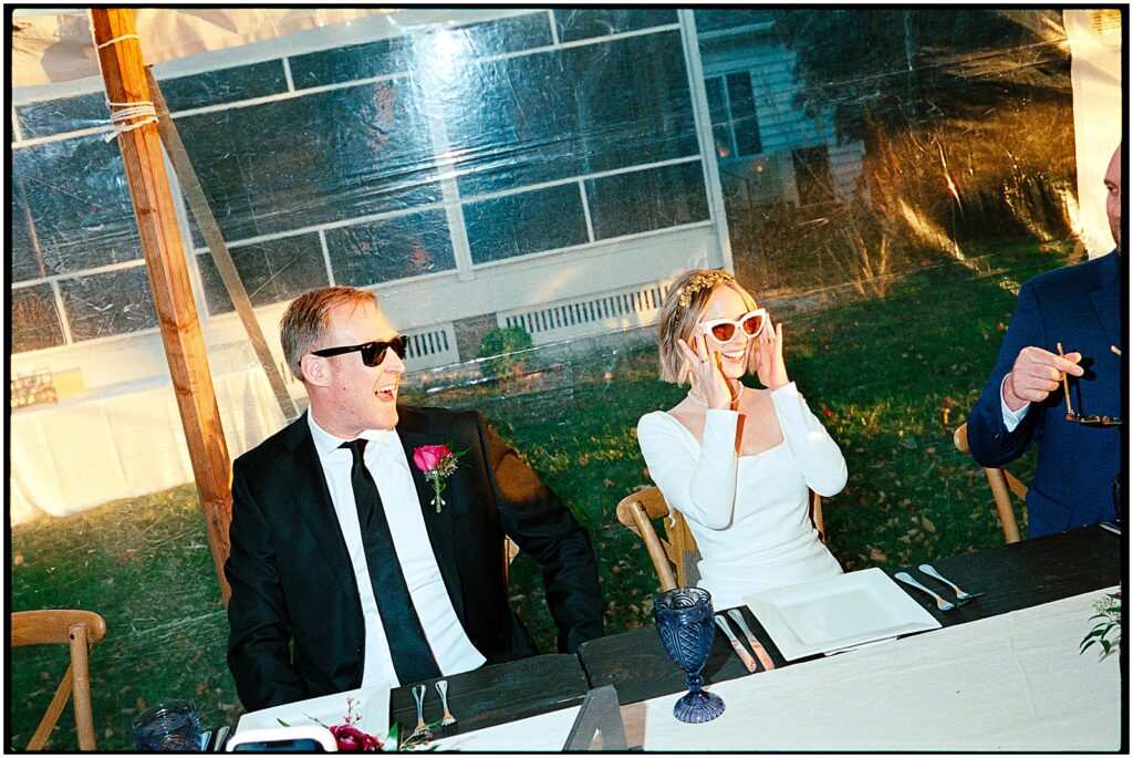 A bride and groom sit at a reception table and put on sunglasses at their lake house wedding.