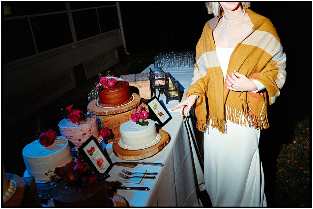 A bride wearing a blanket around her shoulders points to a table of wedding cakes.