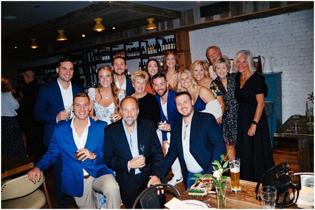 A bride and groom pose with rehearsal dinner guests for a group photo in a Philadelphia restaurant.