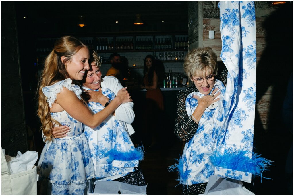 The mother of the groom holds up blue and white pajamas while the bride hugs her mother.