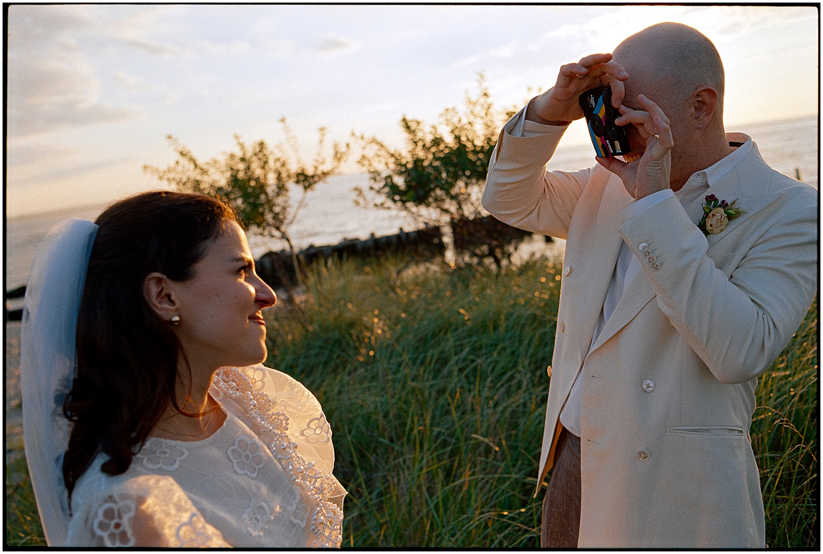 A groom holds up a film camera to take a picture of a bride at their Sandy Hook wedding.