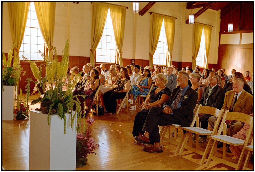 Guests sit in wooden chairs inside the Sandy Hook Chapel watching a wedding ceremony.