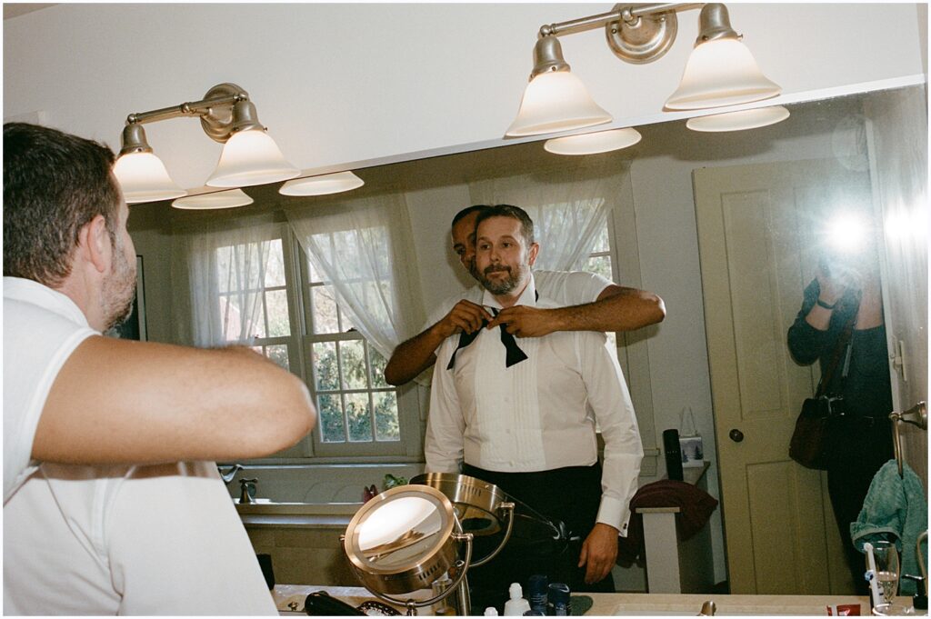 A groomsman helps a groom tie a bowtie in a getting ready suite at Winterthur.