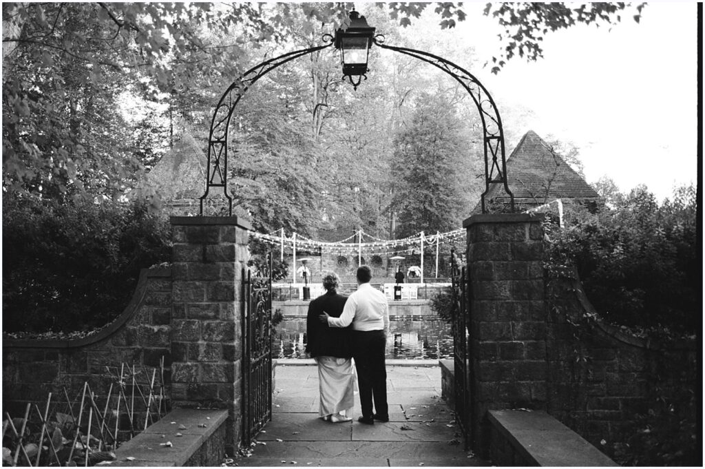 A groom puts his arm around a bride at the entrance of a garden.