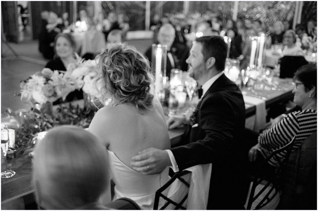 A groom puts his arm around a bride's back at their reception table.