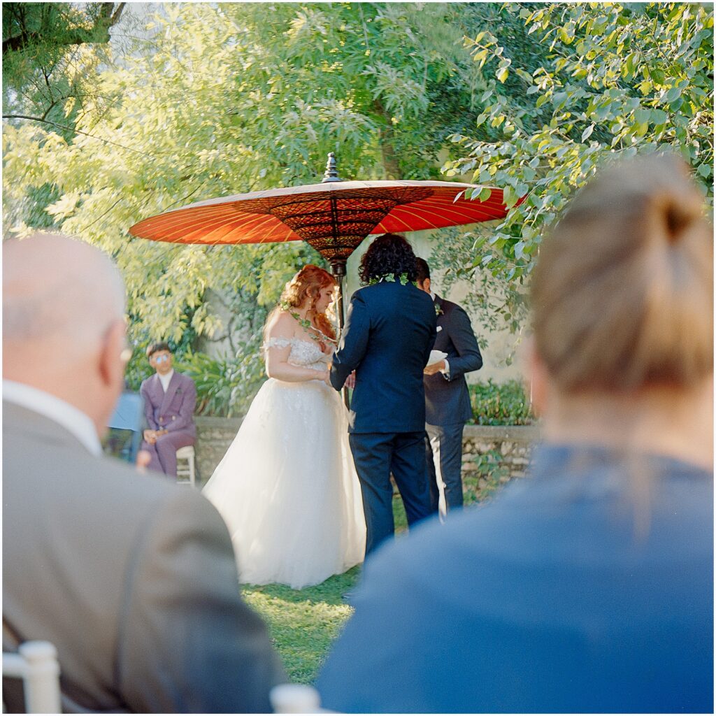Destination wedding photography shows a couple exchanging vows under a large parasol at an Italian villa.
