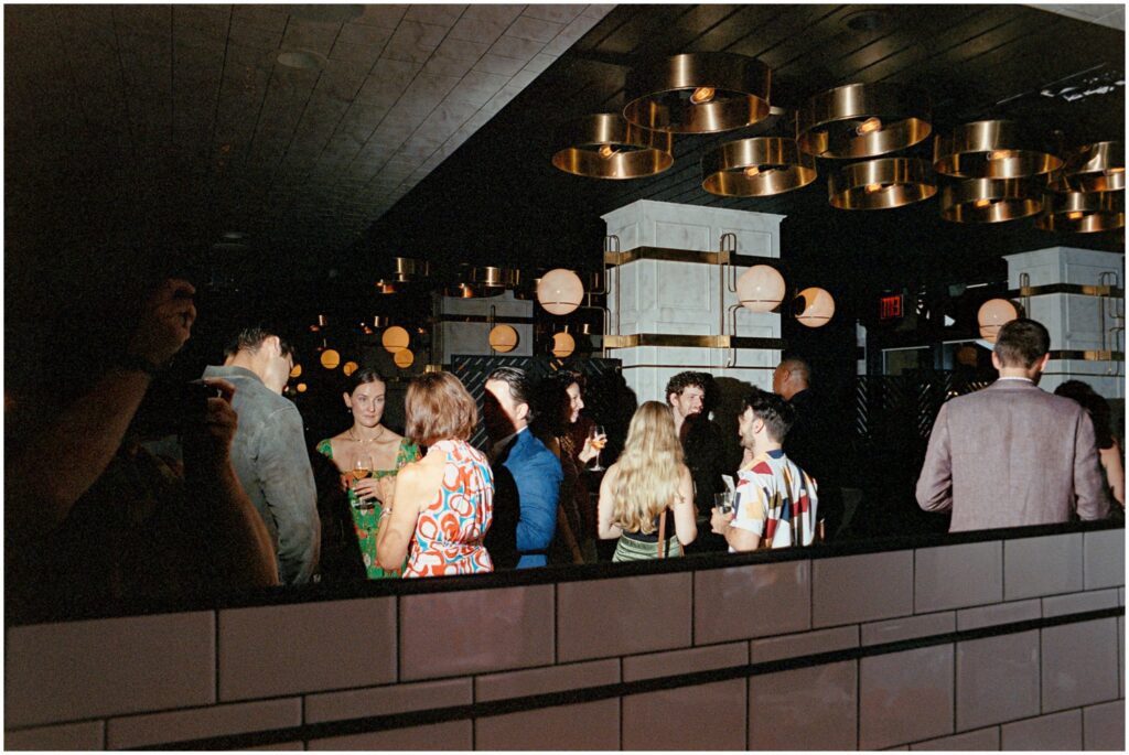 A group of guests, dressed elegantly, mingle and laugh in the chic interior of American Brass, Long Island City, NY, during an engagement party.