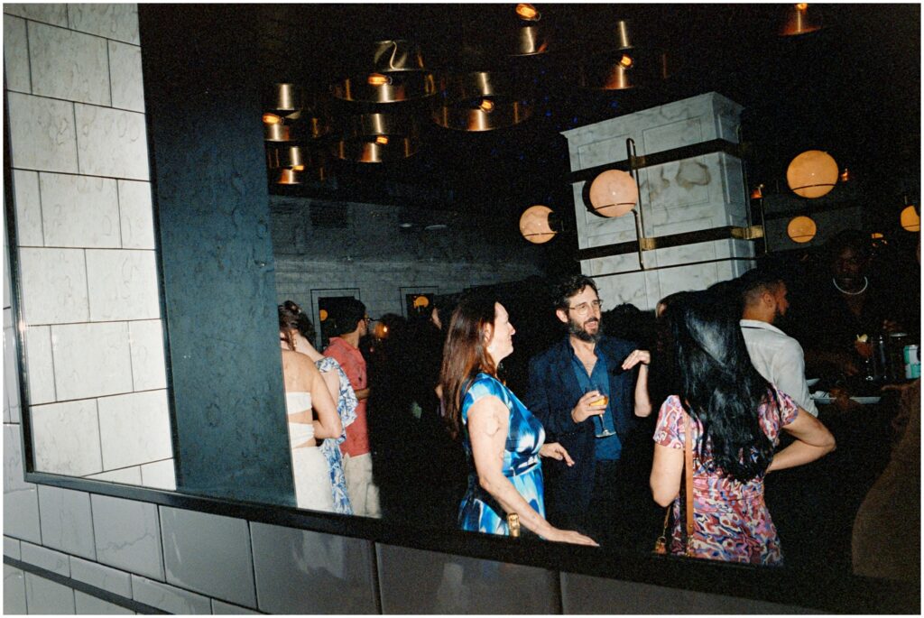 A group of guests, dressed elegantly, mingle and laugh in the chic interior of American Brass, Long Island City, NY, during an engagement party.