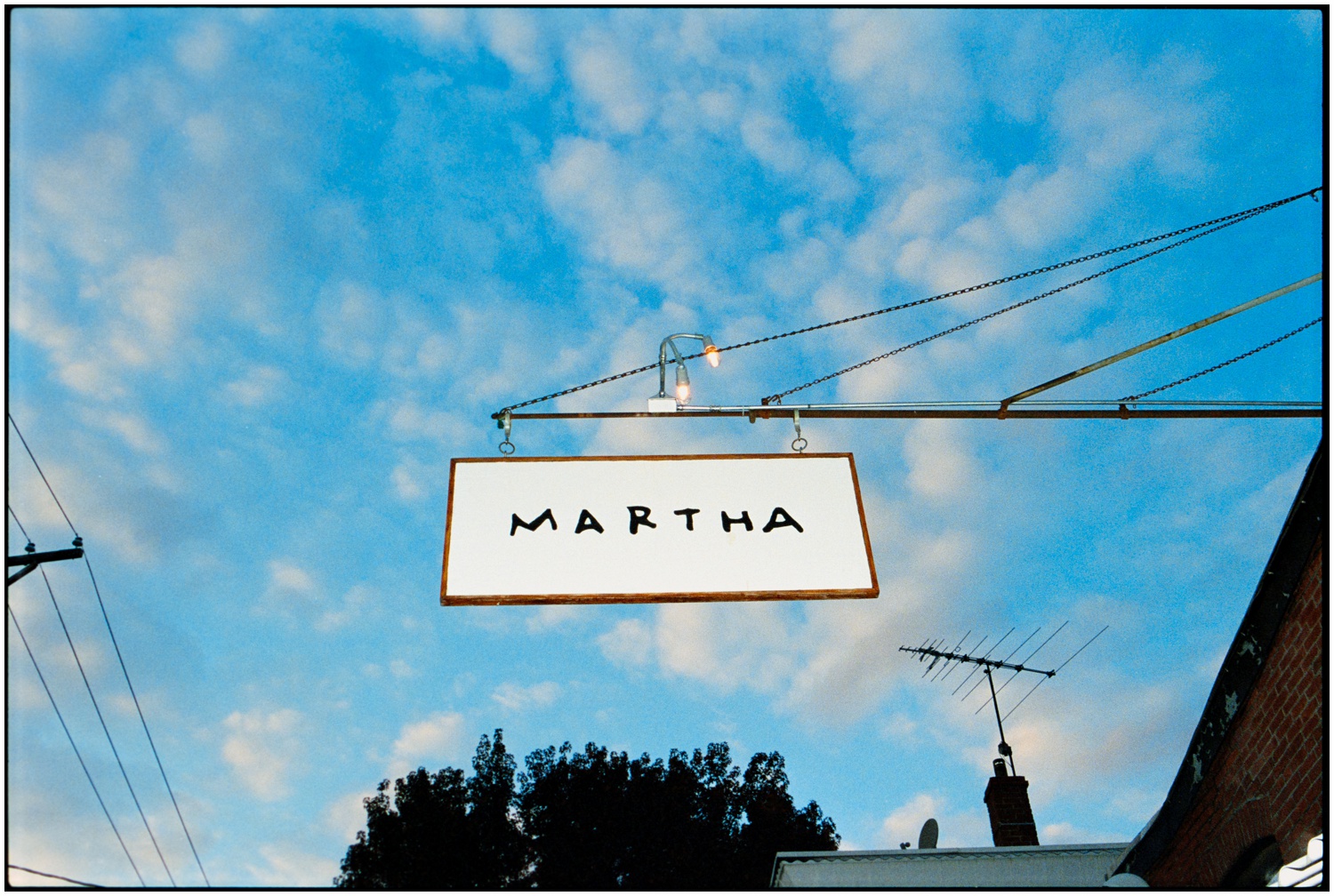 A sign with the text "MARTHA" hangs against a sky with scattered clouds, mounted on a metal arm extending from a brick building in Philadelphia.